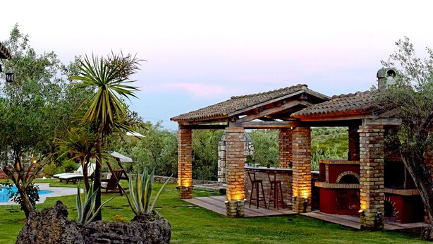 A patio with a brick barbecue and seating area, surrounded by greenery and a pool. - Beautiful 9-Bed Villa in Gouvia with Tennis Court (danilia)