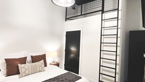 Ein Schlafzimmer mit einem Bett, Kissen, einem Nachttisch mit Lampe, einer Leiter und einem Fenster.