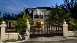 A two-story house with a gated entrance, surrounded by palm trees and a clear sky. - Spacious 3-Bedroom Apartment with WiFi & AC in Lovely Nassau (Nassau)