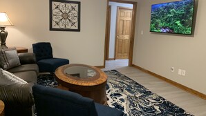 Un salon avec un canapé bleu, une table basse ronde en bois et un tapis à motifs. Un tableau encadré est accroché au mur, et un téléviseur diffuse une scène de la nature.