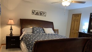 Une chambre à coucher avec un lit en bois, une table de chevet avec une lampe, un ventilateur de plafond, une porte et une œuvre d’art accrochée au mur.
