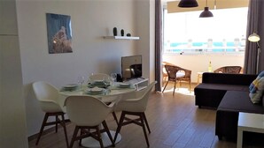 Uma área de jantar com uma mesa posta para quatro pessoas, uma televisão de tela plana e vista para a varanda com móveis de exterior.