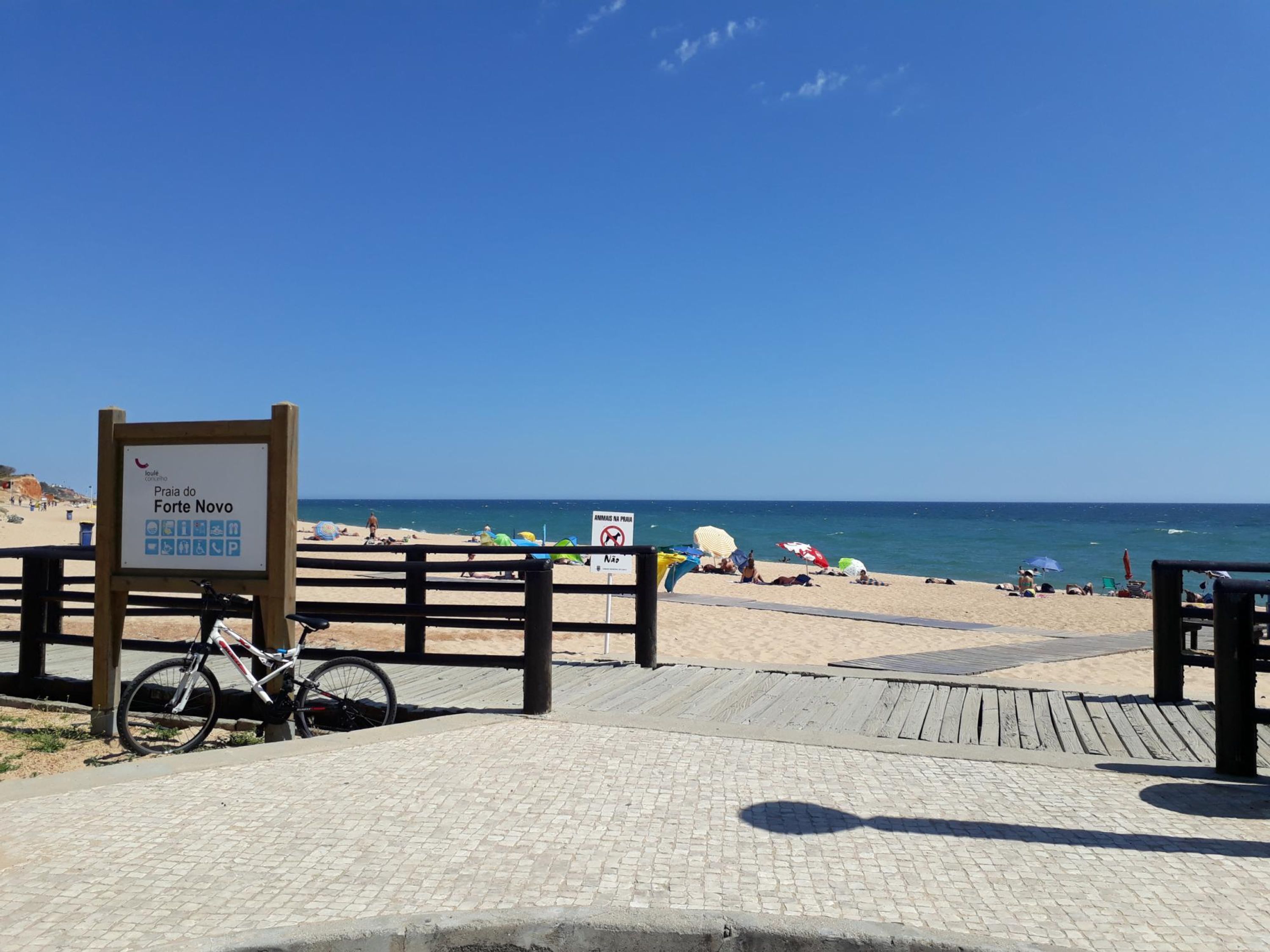 Una platja amb un passeig marítim de fusta, un senyal que indica Praia do Forte Novo, i una bicicleta aparcada al passeig.