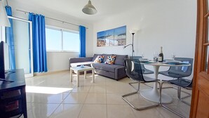 A modern living room with a grey sofa, a coffee table, and a dining area with a table and chairs. - T1 Colina Ferias 8C A C Garagem Vista MAR (Loulé)