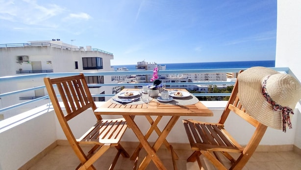 A balcony with a wooden table and chairs, overlooking a coastal cityscape and the sea. - T1 Colina Ferias 8C A C Garagem Vista MAR (Loulé)