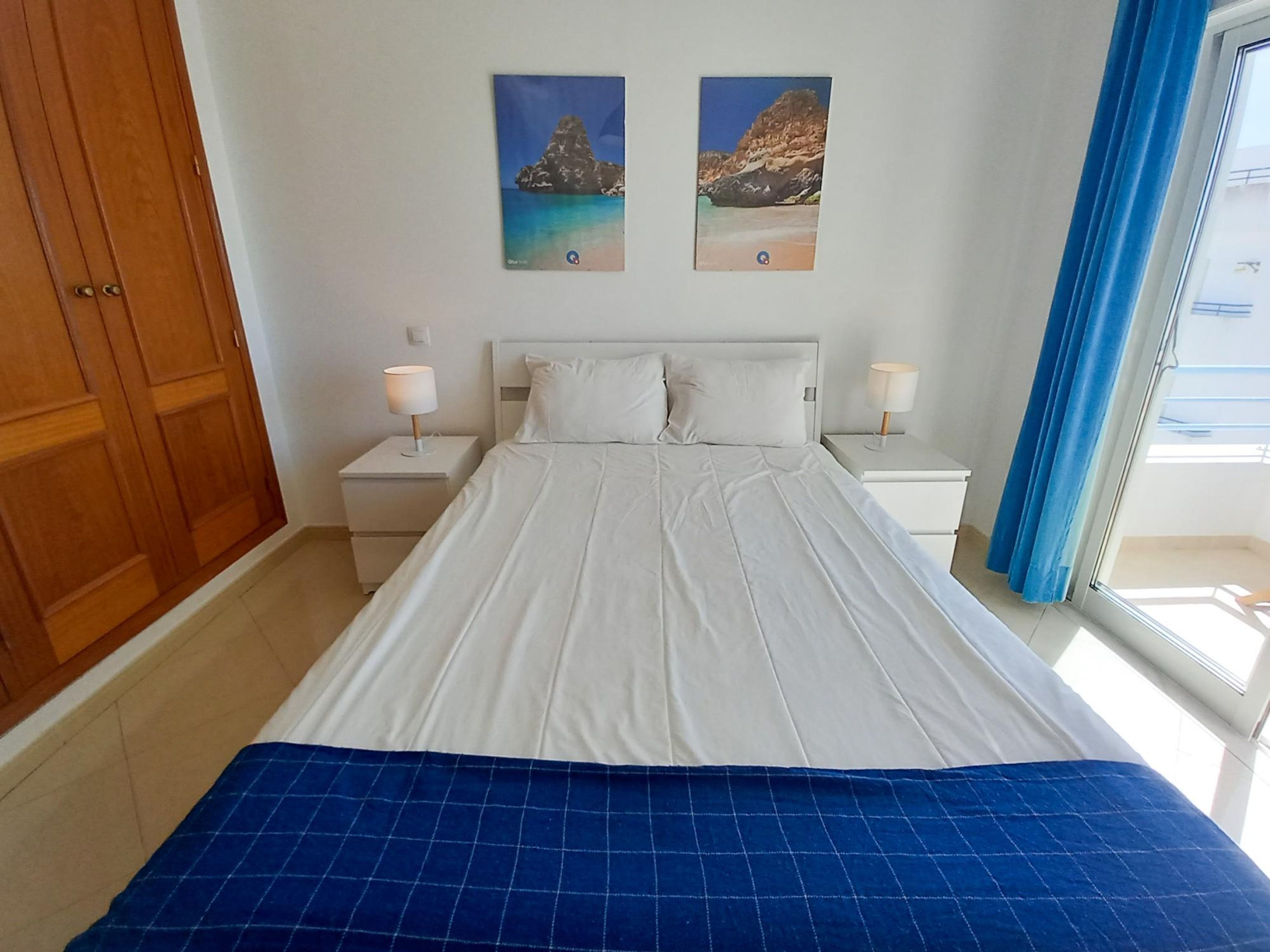 A bedroom with a large bed, two nightstands, a wooden wardrobe, and two wall-mounted paintings of coastal scenes.