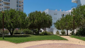 A modern building with a flat roof and balconies, surrounded by a well-maintained garden with trees and a paved walkway. - T1 Colina Ferias 8C A C Garagem Vista MAR (Loulé)