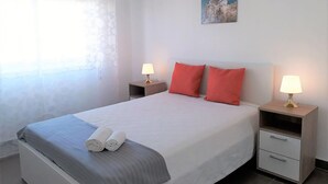 A neatly made bed with pillows, two bedside tables with lamps, a painting on the wall, and a window with curtains. - T1 Torre 20 6G M Gico 80M Praia Wi-fi 4 Pessoas (Loulé)