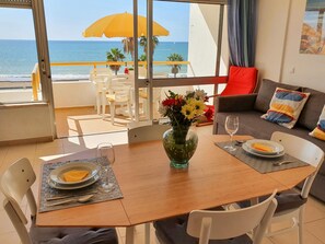 A dining area with a table set for two, a vase with flowers, and a view of the ocean through a balcony.