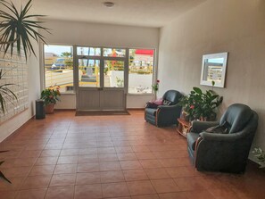A spacious lobby with a tiled floor, a reception desk, and a large glass door leading outside. There are several chairs and potted plants arranged for guests.