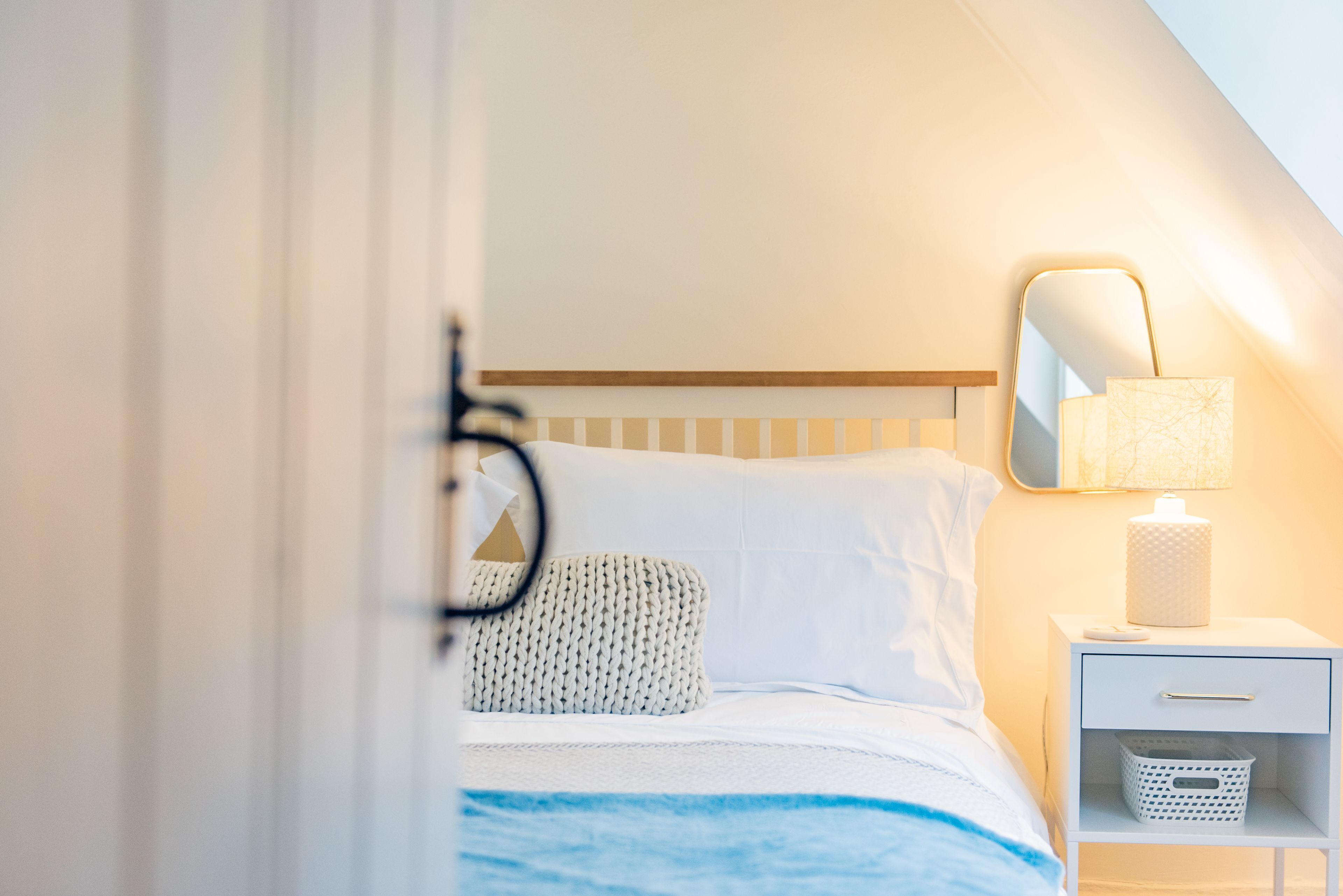 Une chambre à coucher avec un lit, une table de chevet avec une lampe, un petit panier placé sur la table de chevet et un miroir accroché au mur.