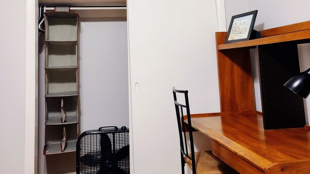 Ein kompakter Raum mit einem Holztisch, einem Stuhl, einer schwarzen Lampe und einer weißen Tür zu einem Schrank mit Regalen und einer Kleiderstange.