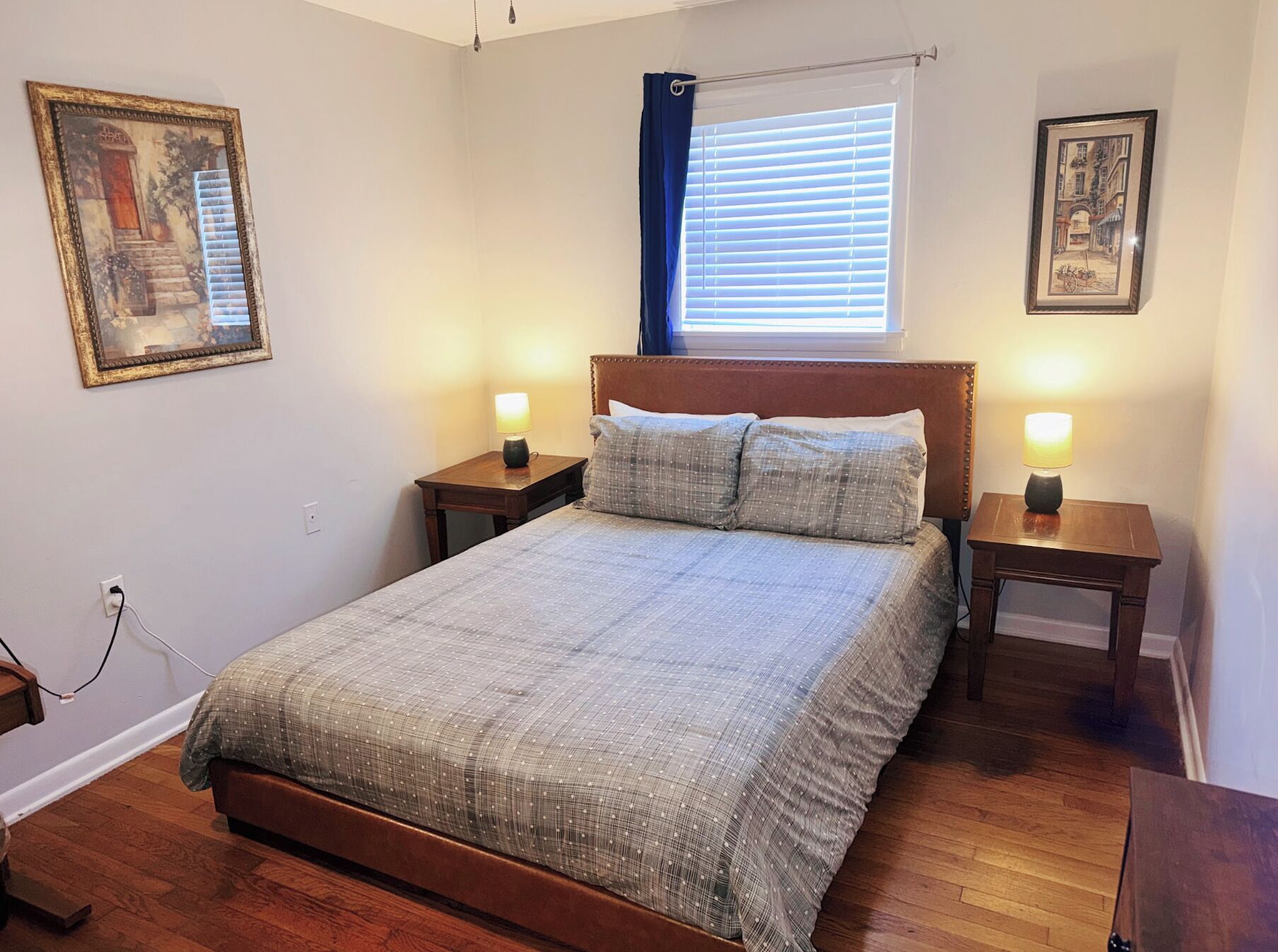 Une chambre à coucher avec un lit, deux tables de chevet, une fenêtre avec des stores et deux tableaux encadrés accrochés aux murs.