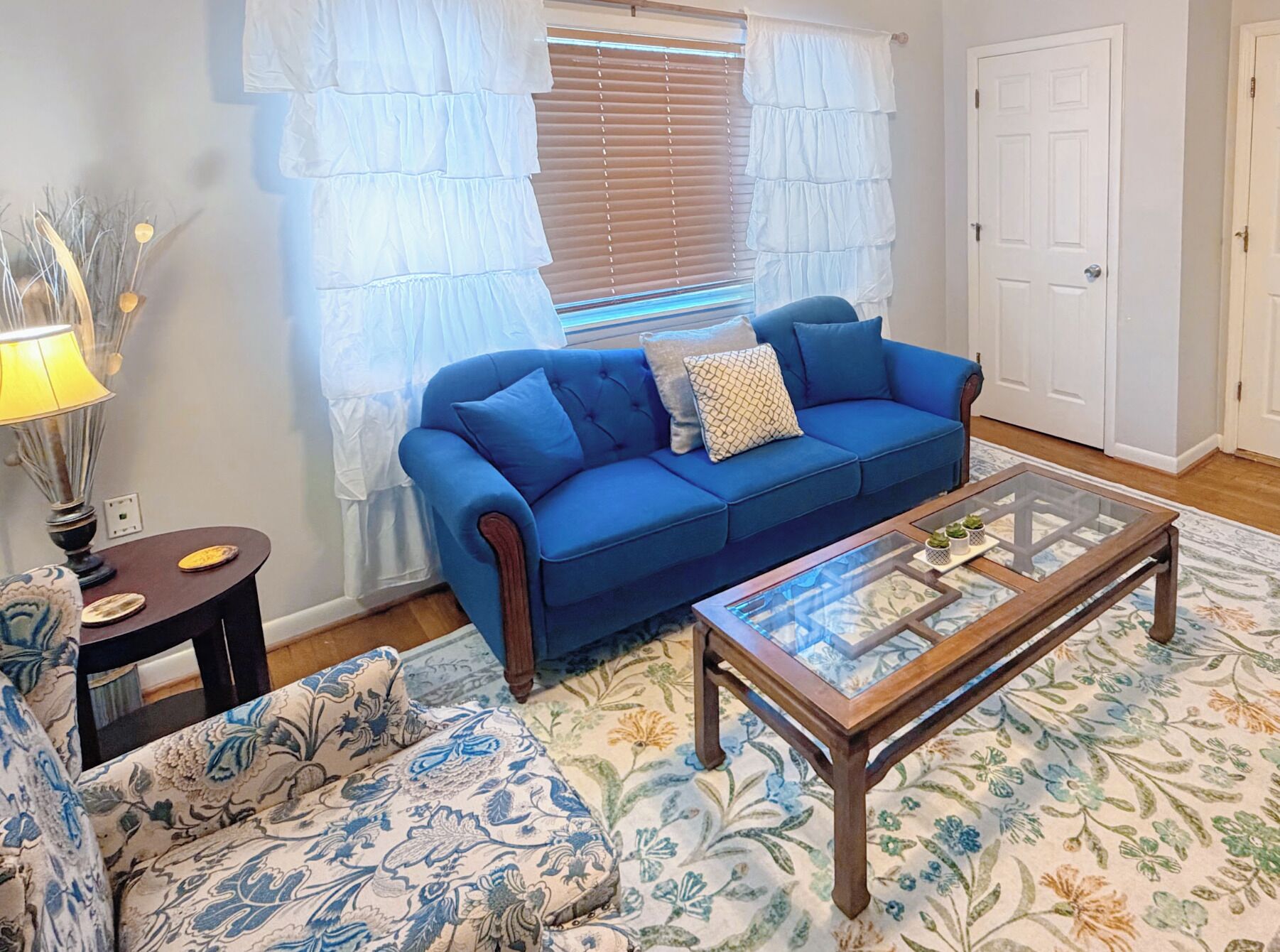 A living room with a blue sofa, a glass coffee table, a patterned rug, and a lamp.