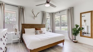 Ein Schlafzimmer mit einem hölzernen Bett, einem Nachttisch, einer Kommode, einem Deckenventilator und einem großen Fenster mit Vorhängen.