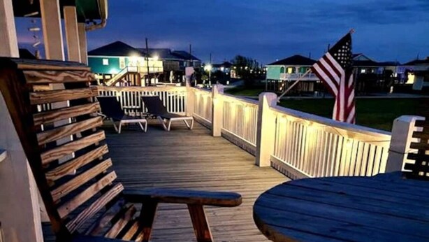 Une terrasse en bois avec des sièges d’extérieur et une rambarde, donnant sur un quartier résidentiel avec des maisons et un drapeau américain.