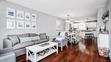 Een moderne woonkamer met een grijze bank, een witte salontafel en grijze fauteuils. Op de achtergrond is een eethoek met een witte tafel en stoelen zichtbaar.