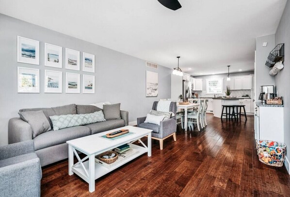 A modern living room with a grey sofa, white coffee table, and grey armchairs. A dining area with a white table and chairs is visible in the background.