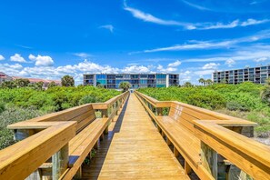 A wooden boardwalk leading to a modern building with multiple floors and balconies, surrounded by lush greenery.