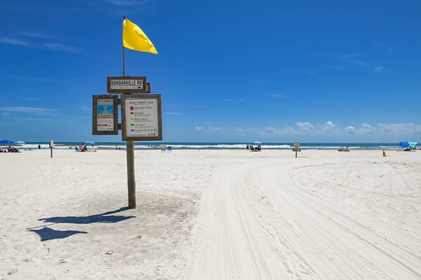 A beach with a signpost indicating 'Dondanville Rd' and 'Beach Warning Flags'.