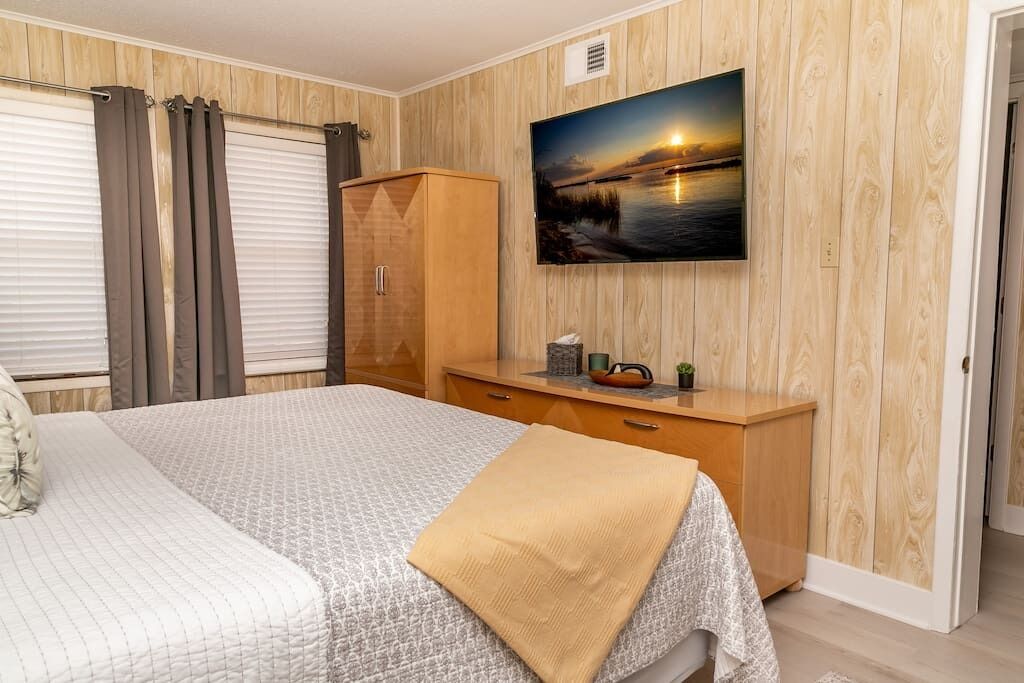 A bedroom with a bed, a wooden wardrobe, a bedside table with a lamp, and a wall-mounted TV displaying a sunset scene.