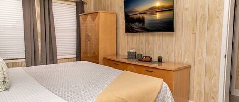 A bedroom with a bed, a wooden wardrobe, a bedside table with a lamp, and a wall-mounted TV displaying a sunset scene.