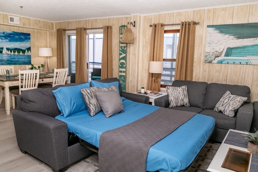 A hotel room with a large bed, a sofa, a desk, and a chair. There are curtains, a lamp, and a painting on the wall.