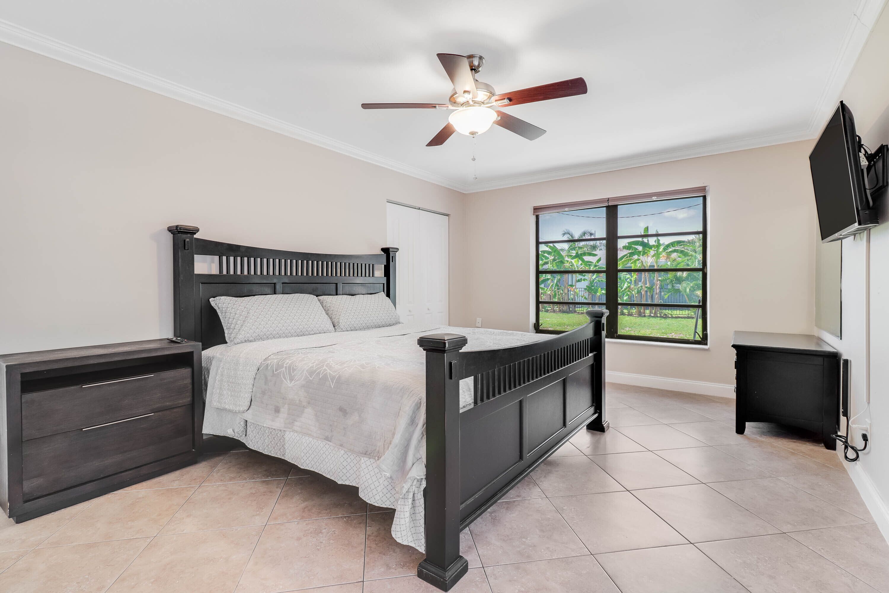 A bedroom with a bed, a nightstand, a dresser, a ceiling fan, a TV, and a window with a view of greenery.