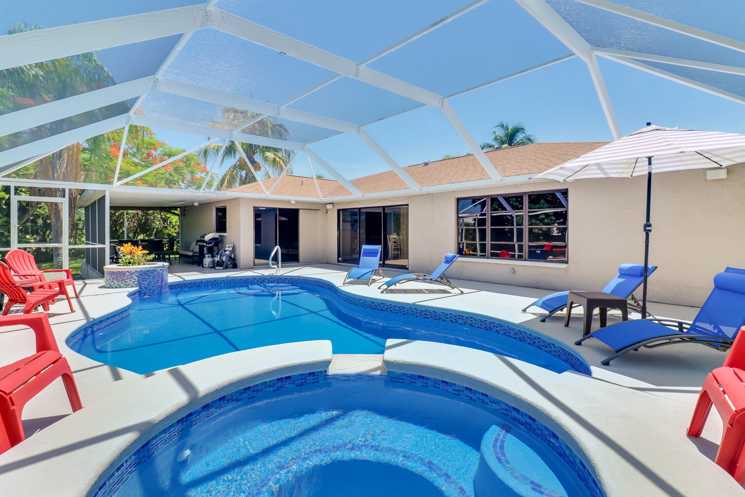A swimming pool with lounge chairs and umbrellas, surrounded by a covered area with a view of palm trees.