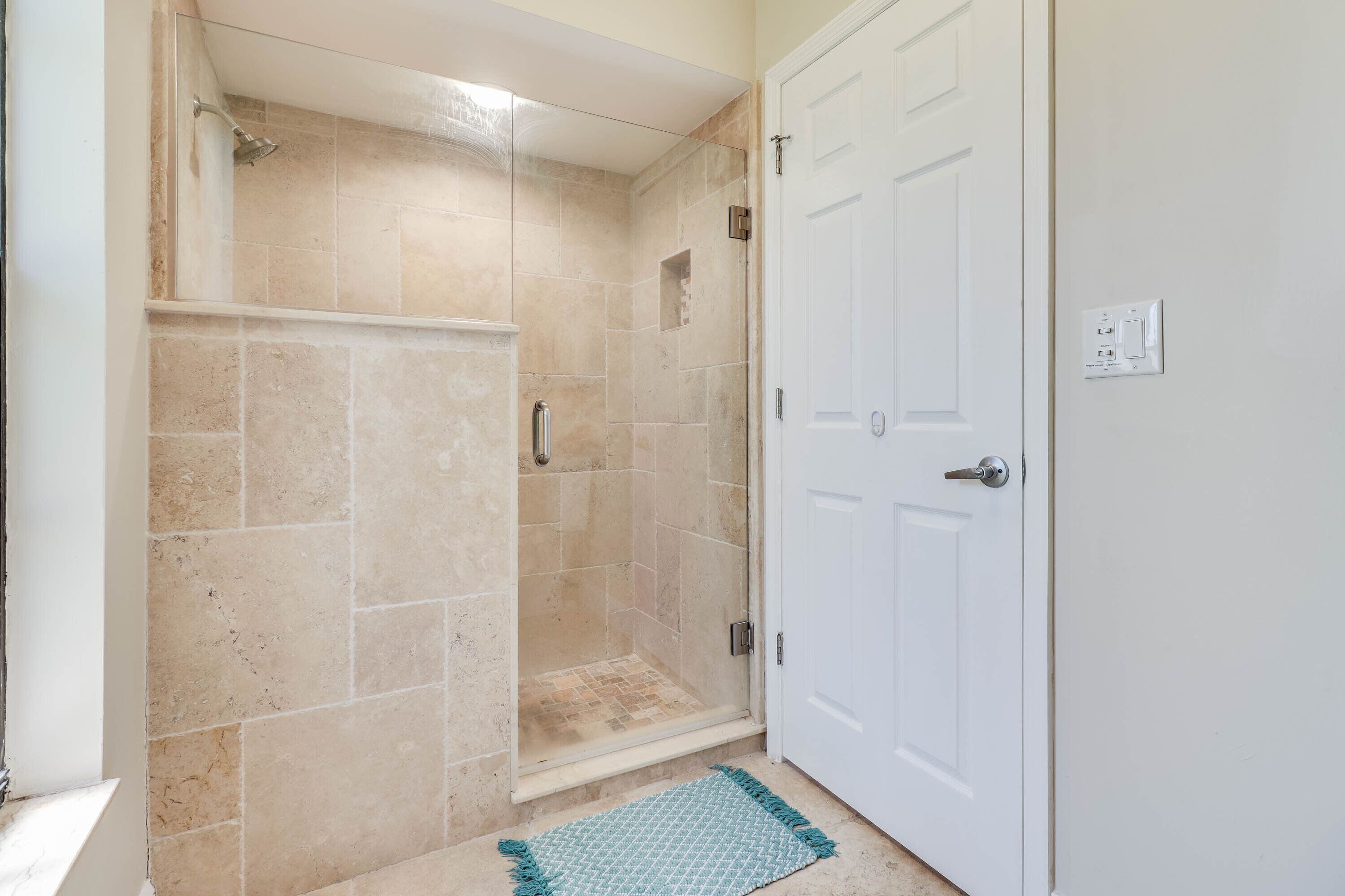 Une salle de bain avec une douche à parois de verre, des carreaux de couleur beige, une porte blanche et un petit tapis sur le sol.