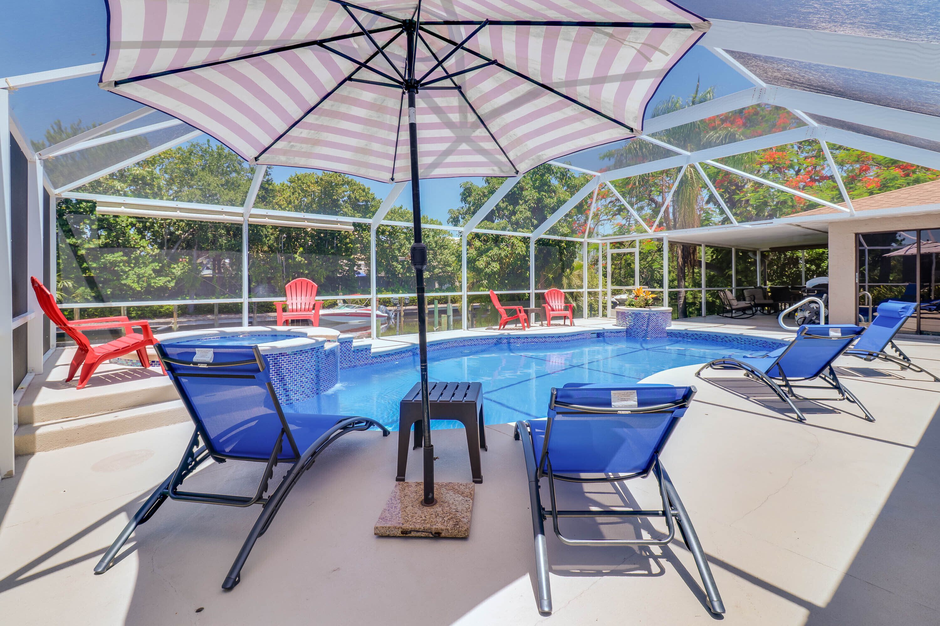 A covered outdoor swimming pool area with blue lounge chairs, a red chair, and a striped umbrella.