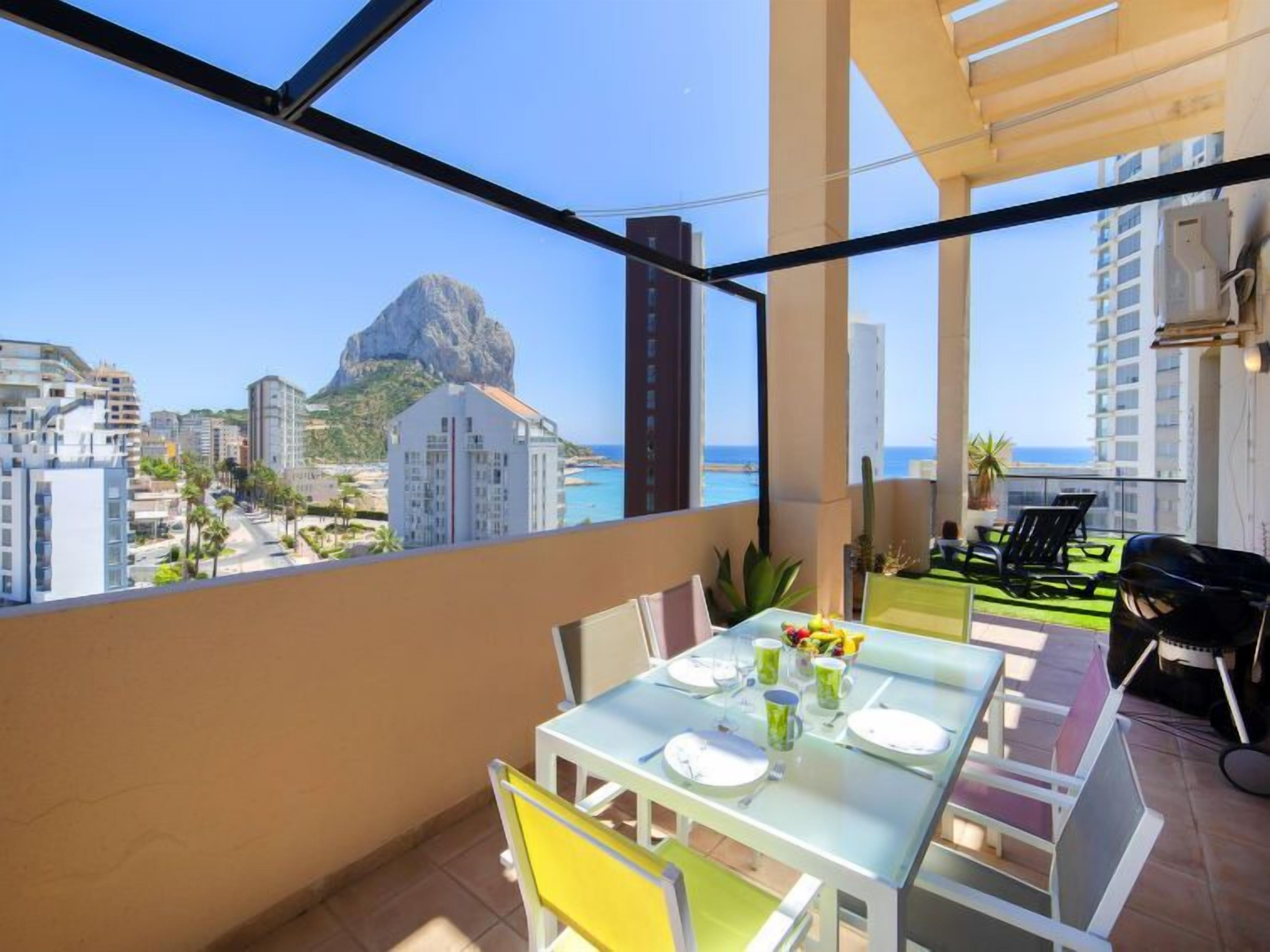 A balcony with a table set for dining, overlooking a coastal cityscape with a prominent mountain.
