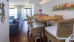 Un espace repas moderne avec une table en bois et des chaises en osier, un arrangement floral central composé d’une orchidée violette et un grand tableau abstrait accroché au mur.