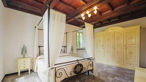 Uma cama com dossel e cortina, teto de madeira, piso de mármore, mesa de cabeceira com castiçal e guarda-roupa amarelo.