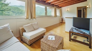 Um quarto espaçoso com teto de madeira, uma janela grande com cortinas, um sofá, uma televisão sobre um suporte de madeira e uma mesinha com uma revista.