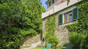 Uma casa de pedra com escadaria de pedra que leva a um deck de madeira. Há duas poltronas de jardim verdes no deck.