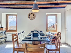 Une salle à manger avec une longue table, des chaises, une horloge et des fenêtres donnant sur une vue sur la montagne.