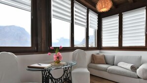 Une pièce chaleureuse avec un plafond en bois, un canapé blanc, une table en verre ornée d’un vase de fleurs et de grandes fenêtres équipées de stores.