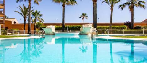 A clear blue pool with lounge chairs, palm trees, and a building in the background.
