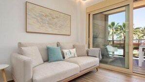Una sala moderna con un sofá color beige, una mesita auxiliar y un ventanal con vistas a palmeras y una piscina.