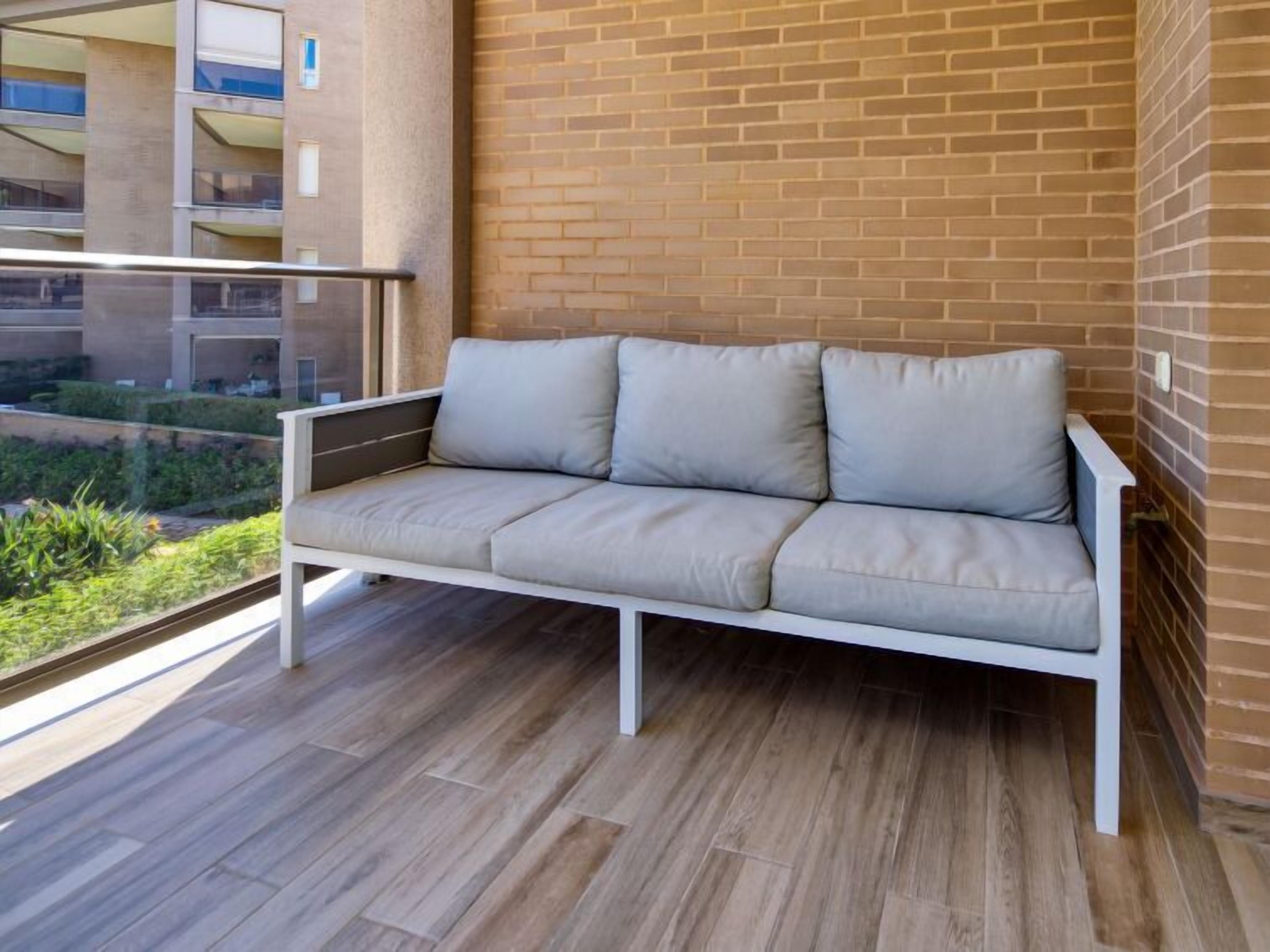 A modern balcony with a white sofa and a view of a brick building and greenery.