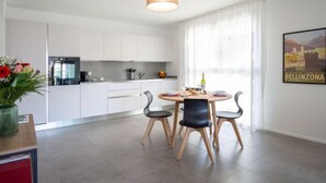 A modern kitchen with white cabinets, a dining area with a round table and four chairs, and a large window with sheer curtains. - LocTowers A3.4.3 by Interhome (Locarno)