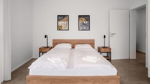 Une chambre moderne avec un lit en bois, du linge de lit blanc, deux œuvres d’art accrochées au mur et des tables de chevet avec des lampes.