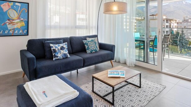 A modern living room with a dark blue sofa, patterned cushions, a coffee table, and a large window with a view of buildings and mountains. - LocTowers A3.3.1 by Interhome (Locarno)