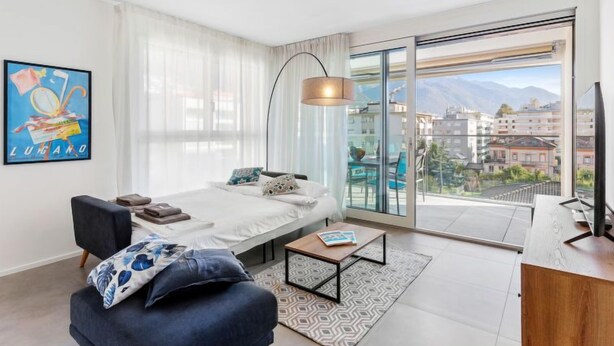 A modern bedroom with a bed, a desk, and a chair. There is a large window with a view of buildings and mountains. - LocTowers A4.2.3 by Interhome (Locarno)