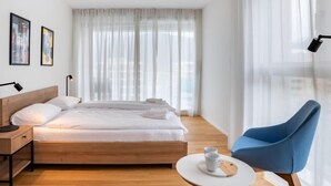 Una camera d'albergo moderna con un letto di legno, un tavolino con due tazze, una sedia blu e un'ampia finestra con tende leggere.
