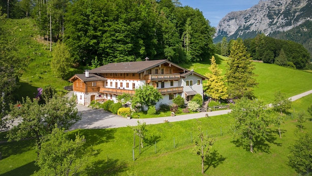 A two-story wooden house with a balcony, surrounded by lush green fields and trees, with mountains in the background.