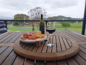 Une table en bois sur laquelle sont disposés un plateau de fromages, des crackers et une bouteille de vin, le tout sur un balcon offrant une vue sur une route et des arbres.