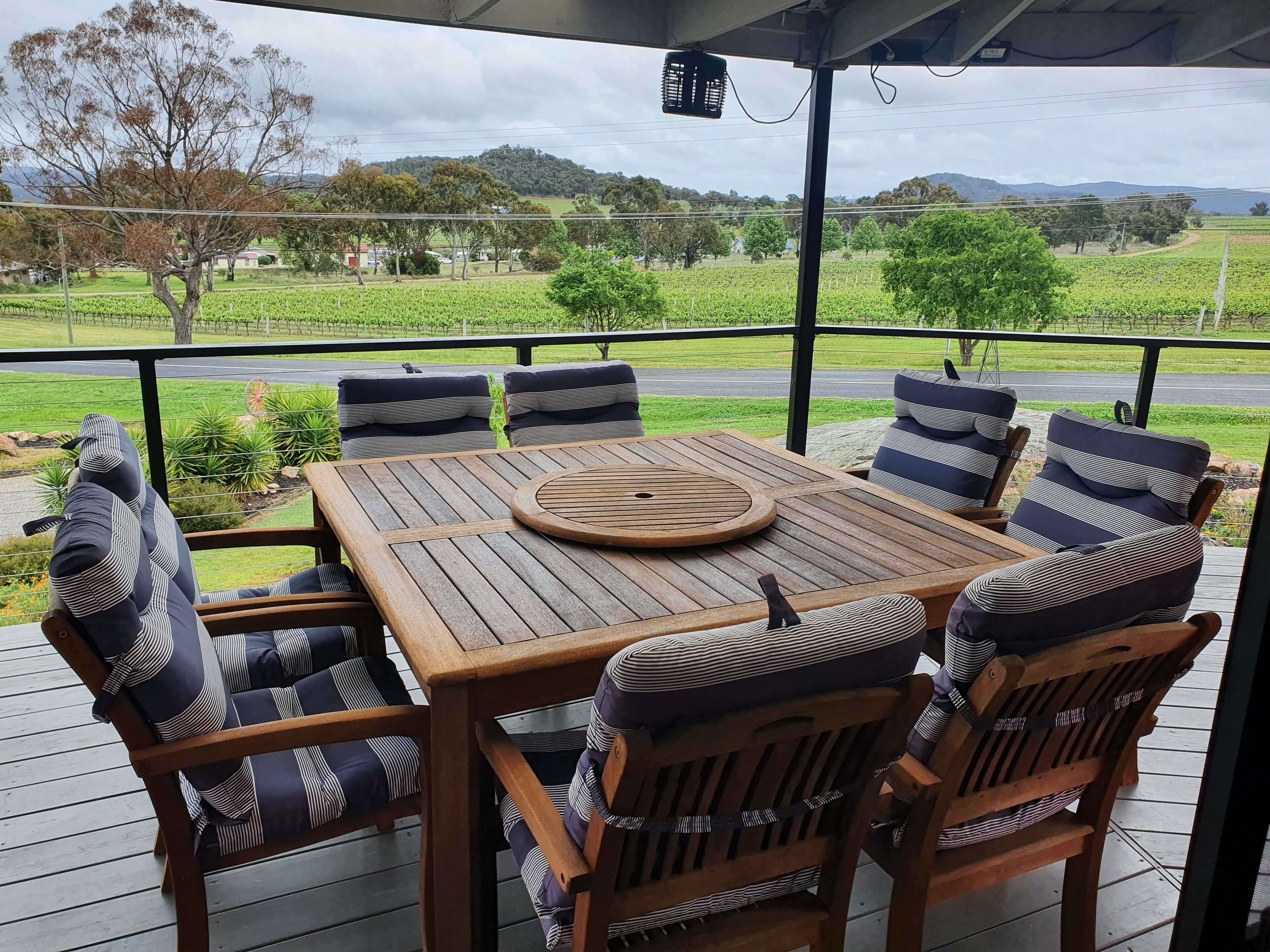 Un espacio de estar al aire libre cubierto con una mesa de madera y sillas acolchadas a rayas, con vistas a un viñedo y una carretera.