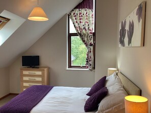 Une chambre avec un plafond en pente, un lit avec du linge de lit blanc et des coussins violets, une table de chevet avec une lampe, un téléviseur posé sur une commode en bois et une fenêtre ornée de rideaux à motifs floraux.